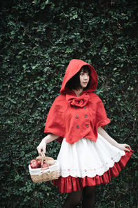 Woman in little red riding hood costume holding basket while standing against plants