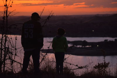 Silhouette man and child standing by lake against sky during sunset