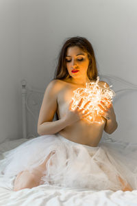 Sensuous young woman covering breasts with illuminated string lights on bed at home