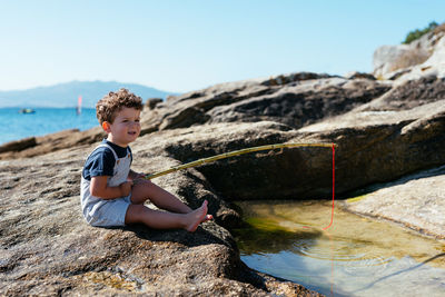 Side view of adorable child fisher in overall holding small rod and fishing in shallow clear brook on rocks