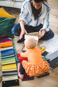 High angle view of female creative professional working while loving daughter playing with fabric swatch on hardwood flo