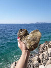 Cropped hand of woman holding heart shape rock against sea