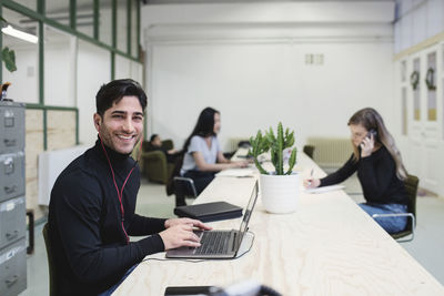 Portrait of happy young man sitting at desk with colleagues working in background