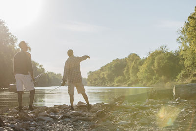 Rear view of male friends with fishing rods standing at lakeshore against sky during sunny day