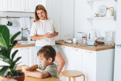 Young woman mother looking as her toddler boy son having breakfast at the table in kitchen at home