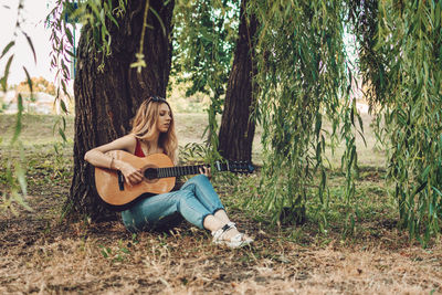 Full length of woman playing guitar while sitting under tree