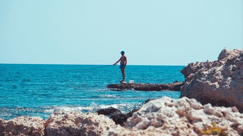 Man fishing while standing on rocks by sea against clear sky