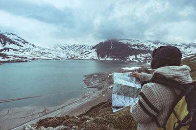 Side view of mature woman holding map while standing at lakeshore against cloudy sky during winter
