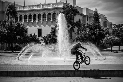 Man riding bicycle by fountain in city against sky