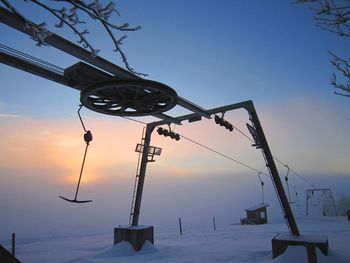 Ski lift cables over snow covered field against sky during sunset