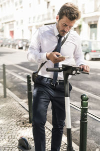 Young businessman with e-scooter using mobile phone in the city, lisbon, portugal