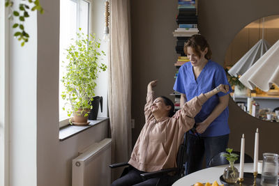 Happy nurse talking to woman stretching arms while sitting on wheelchair at home