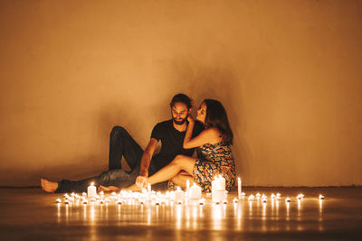Couple sitting by lit candles on floor against wall in darkroom