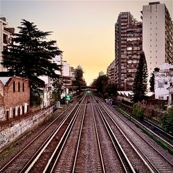 Railroad tracks by buildings against sky at sunset