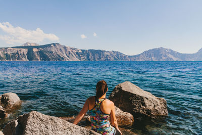 Rear view of woman sitting on rock by sea against mountain