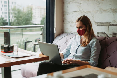 Young woman wearing mask using laptop while sitting at cafe