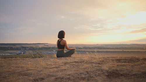 Side view of woman sitting on field against sky during sunset