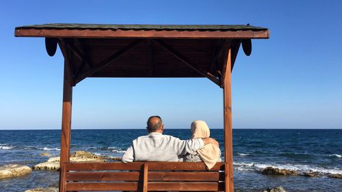 Rear view of couple sitting on beach against clear sky