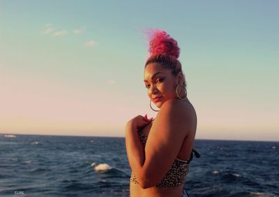 Young woman looking away while standing in sea against sky during sunset