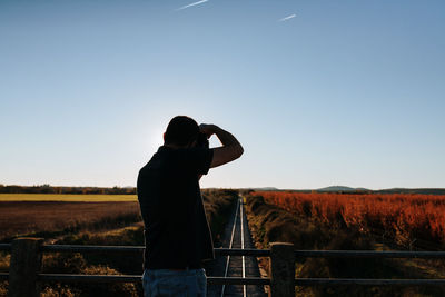 Rear view of man photographing railroad tracks from bridge against sky