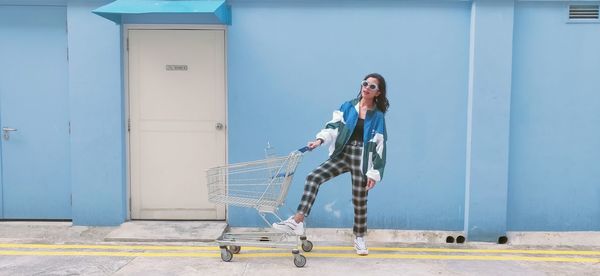Full length of woman wearing sunglasses standing by shopping cart against blue wall