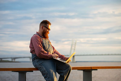 Bearded hipster man freelancer using laptop,  writing message, chatting, shopping at embankment 