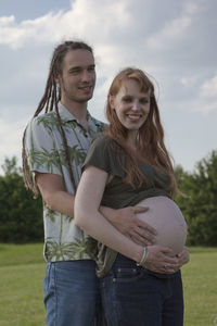 Portrait of happy pregnant woman with man standing on field against sky