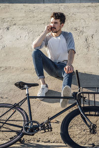 Young man with commuter fixie bike sitting on concrete wall talking on cell phone