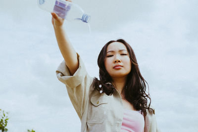 Low angle portrait of confident young woman pouring water from bottle against sky