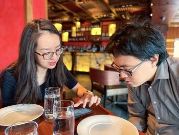 Side view of young asian man and woman in eyeglasses having a discussion over lunch in a restaurant.