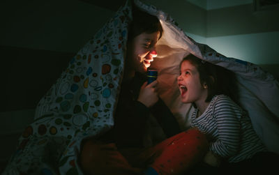 Smiling siblings with flashlight lying down on bed in darkroom