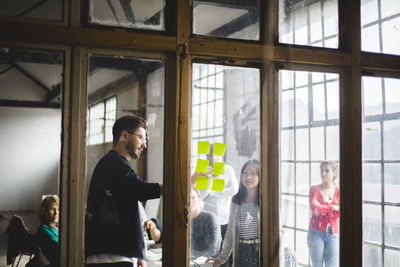 Young male it programmer sharing business ideas with team while pointing at adhesive notes seen through glass wall in of