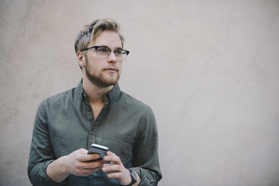 Thoughtful male computer programmer holding smart phone against beige wall in office