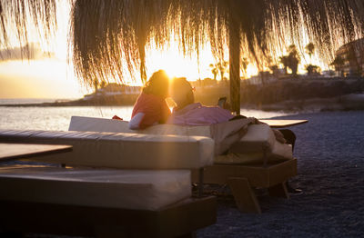 People relaxing on shore by sea against sky during sunset