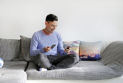 Full length of man using phone while holding credit card on sofa at home