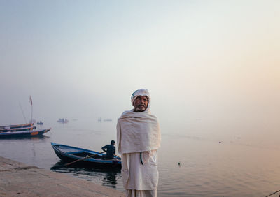 Varanasi, india - february, 2018: elderly indian man wearing traditional white clothes standing on river bank with fishing boats on water in early foggy morning