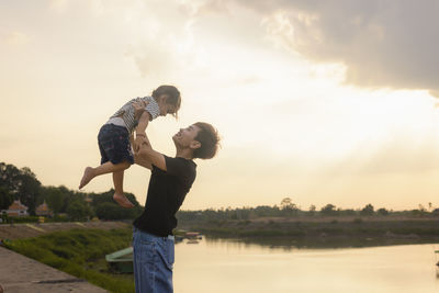 Rear view of woman with arms raised standing against lake during sunset