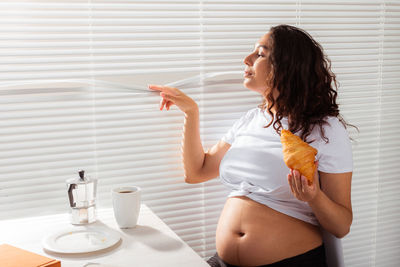 Pregnant woman holding croissant looking through blinds