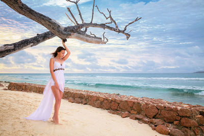 Full length of woman wearing white dress standing at beach against sky