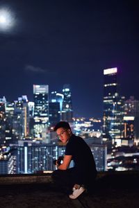 Young man crouching against illuminated cityscape on building terrace at night