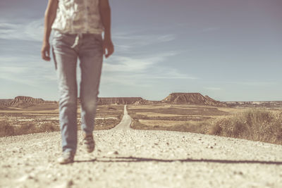 Low section of woman walking on dirt road at desert against blue sky