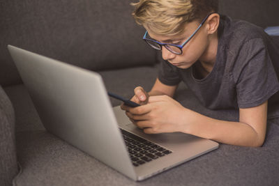 Close-up of boy using phone over laptop while lying on sofa