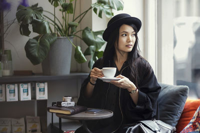 Thoughtful young woman holding coffee cup while sitting by window in creative office