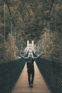 Rear view of man standing with arms outstretched on footbridge in forest