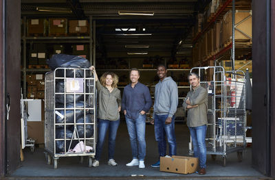 Portrait of smiling coworkers standing amidst carts at warehouse doorway