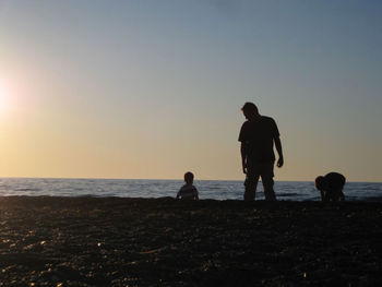 Silhouette father standing by sons at beach against clear sky during sunset