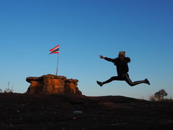 Low angle view of man jumping on rock against clear blue sky