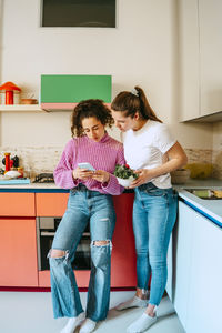 Young woman sharing smart phone with female friend while leaning on kitchen counter at home