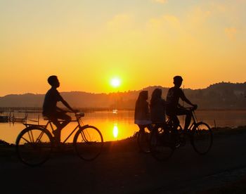 Silhouette people riding bicycles on field by lake against orange sky during sunset