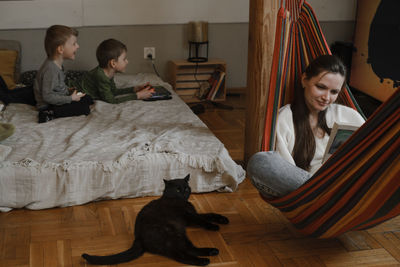 Kids are playing play station while mother is reading book in hammock. family with black cat as a